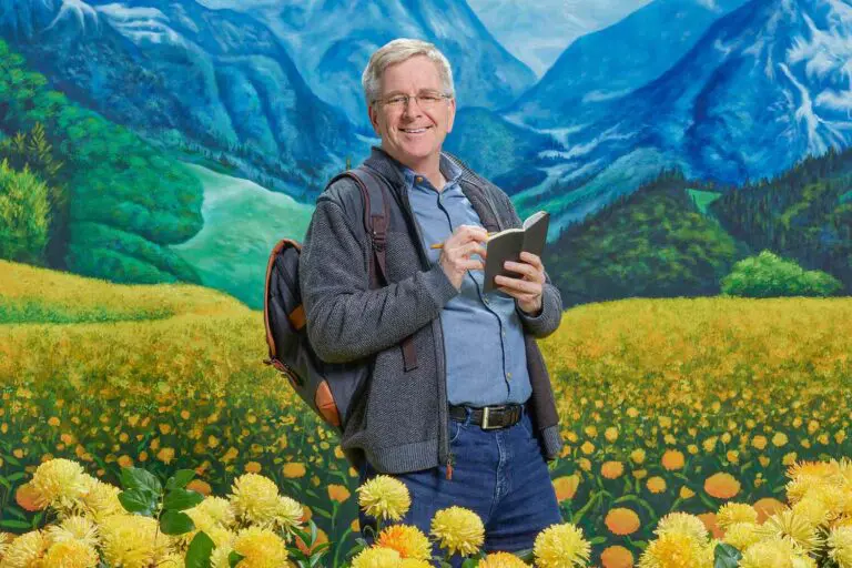 Rick Steves standing in front of a painting of mountains and yellow flowers